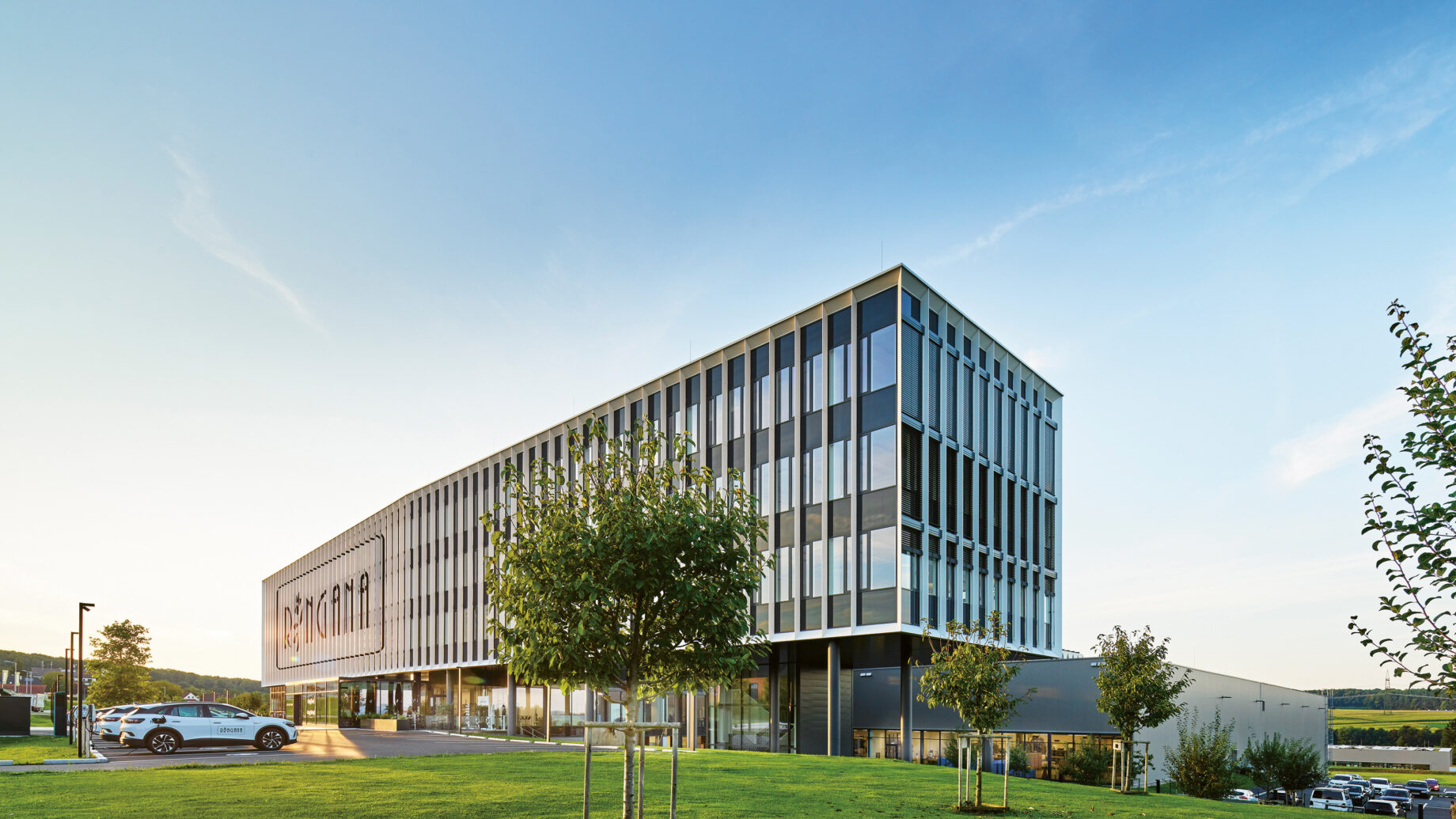 Ein modernes Bürogebäude mit großen Fenstern, umgeben von Rasenflächen und Bäumen, unter einem strahlend blauen Himmel. In der Nähe steht ein weißes Auto.
