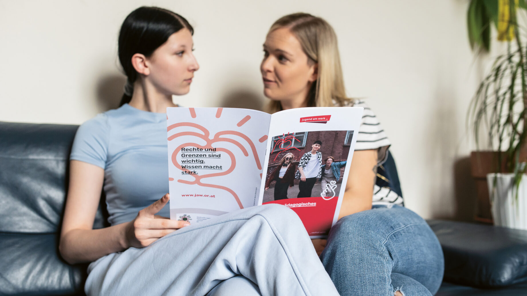 Zwei Frauen sitzen auf einem Sofa, schauen sich an und halten eine Broschüre mit Text und Bildern auf dem Einband in den Händen, die sie gerade durchblättern.