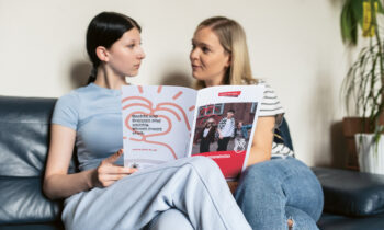 Zwei Frauen sitzen auf einem Sofa, schauen sich an und halten eine Broschüre mit Text und Bildern auf dem Einband in den Händen, die sie gerade durchblättern.