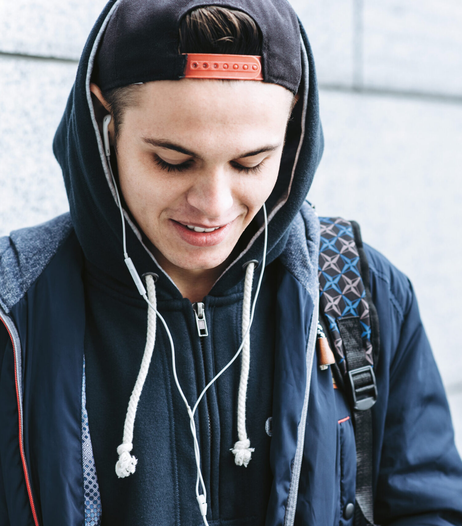 Ein junger Mann mit Kapuzenpulli, Mütze und Rucksack lächelt, während er nach unten schaut und Musik über seine Kopfhörer hört; im Hintergrund ist eine helle Steinmauer zu sehen.