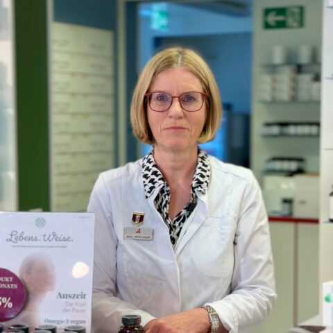 Eine Frau in einem weißen Laborkittel steht in einer Apotheke und lächelt leicht. Im Hintergrund sind Regale mit Flaschen und ein Schild auf Deutsch zu sehen.