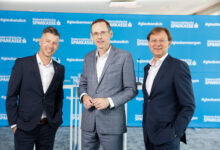 Drei Männer in Anzügen stehen lächelnd vor einer blauen Kulisse der Steiermärkischen Sparkasse im Herzen von Echt GRAZ, der Grazer Innenstadt.