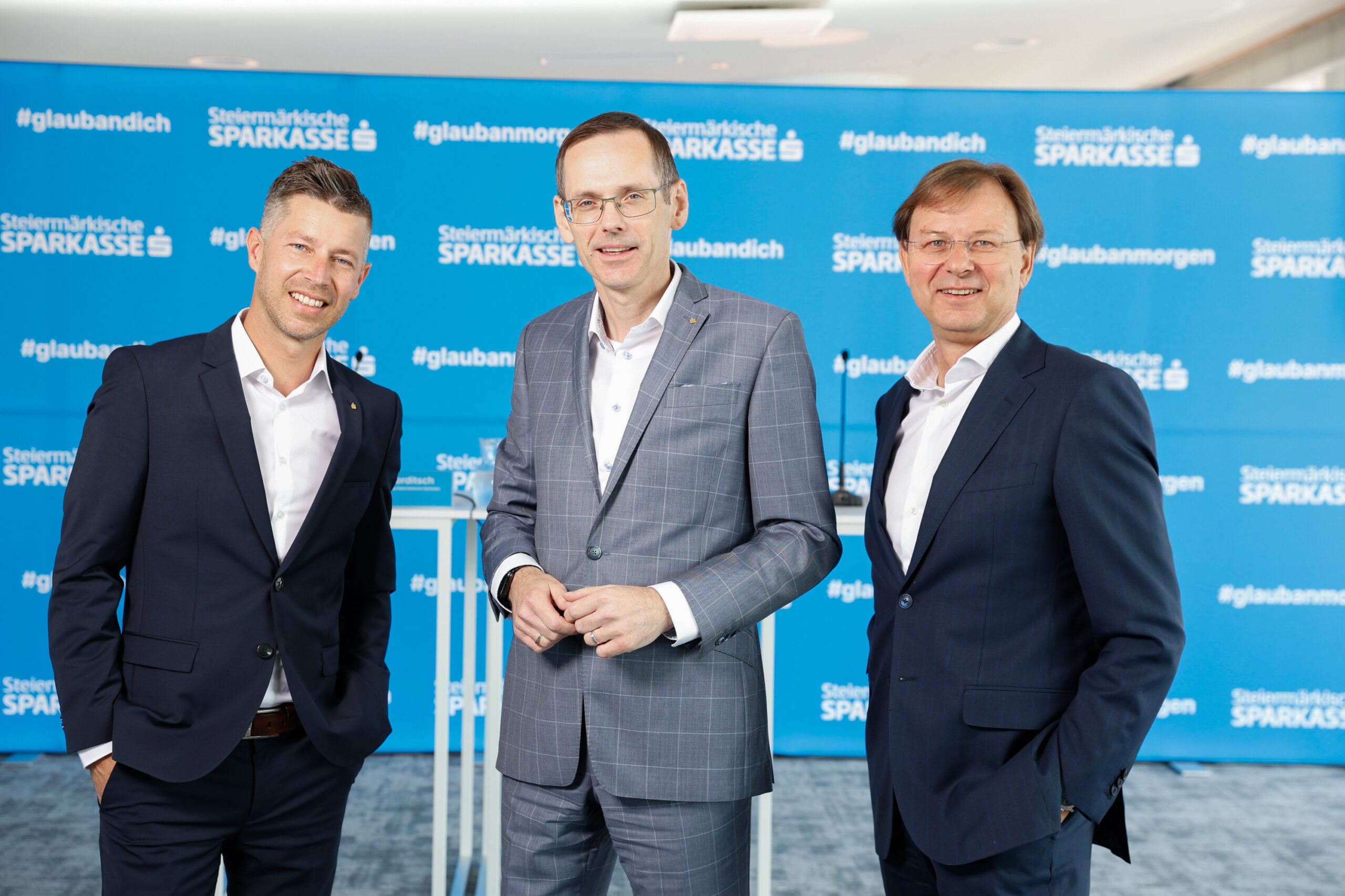 Drei Männer in Anzügen stehen lächelnd vor einer blauen Kulisse der Steiermärkischen Sparkasse im Herzen von Echt GRAZ, der Grazer Innenstadt.