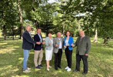 Sechs Erwachsene lächeln und halten Getränke in der Hand, während sie in einem sonnendurchfluteten Park in der Nähe von Echt GRAZ in der Grazer Innenstadt sitzen, umgeben von üppigem grünem Gras und hohen Bäumen.