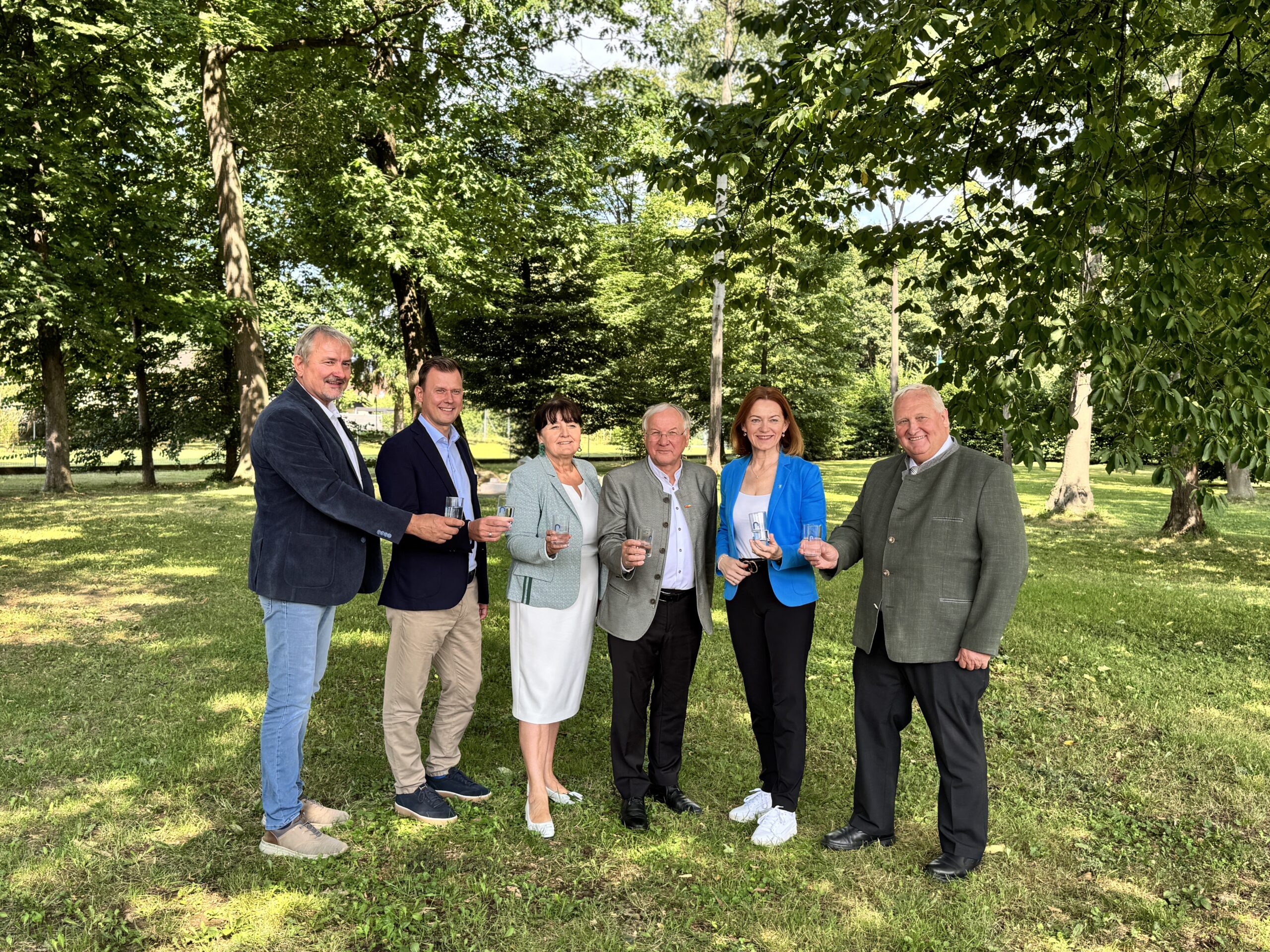 Sechs Erwachsene lächeln und halten Getränke in der Hand, während sie in einem sonnendurchfluteten Park in der Nähe von Echt GRAZ in der Grazer Innenstadt sitzen, umgeben von üppigem grünem Gras und hohen Bäumen.