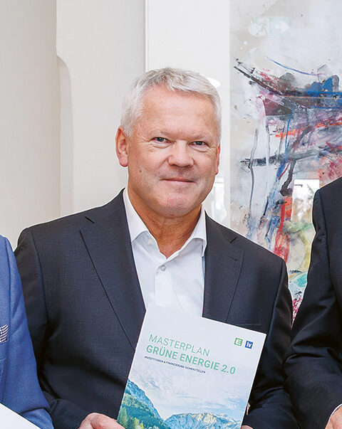 Fünf Männer in Anzügen stehen in einem Raum, lächeln und halten Berichte mit grünen Einbänden in der Hand, auf denen eine Berglandschaft abgebildet ist.