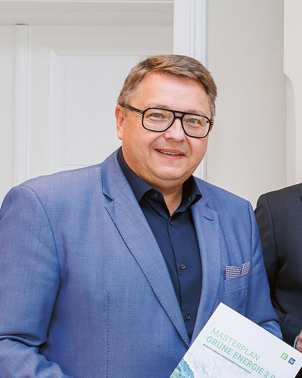 Fünf Männer in Anzügen stehen in einem Raum, lächeln und halten Berichte mit grünen Einbänden in den Händen, auf denen Landschaften abgebildet sind. An der Wand hinter ihnen hängt ein gerahmtes abstraktes Gemälde.