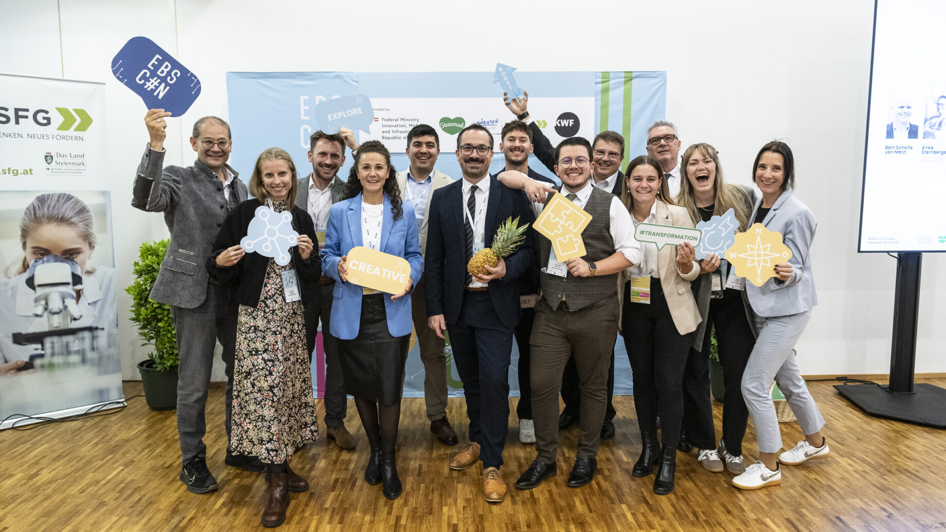 Eine Gruppe beim „Hummelnest Start-up Accelerator Graz“ posiert in einem Innenraum, lächelt und hält motivierende Schilder und Symbole in die Kamera; eine Person hält eine Ananas in der Hand.