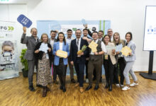 Eine Gruppe beim „Hummelnest Start-up Accelerator Graz“ posiert in einem Innenraum, lächelt und hält motivierende Schilder und Symbole in die Kamera; eine Person hält eine Ananas in der Hand.