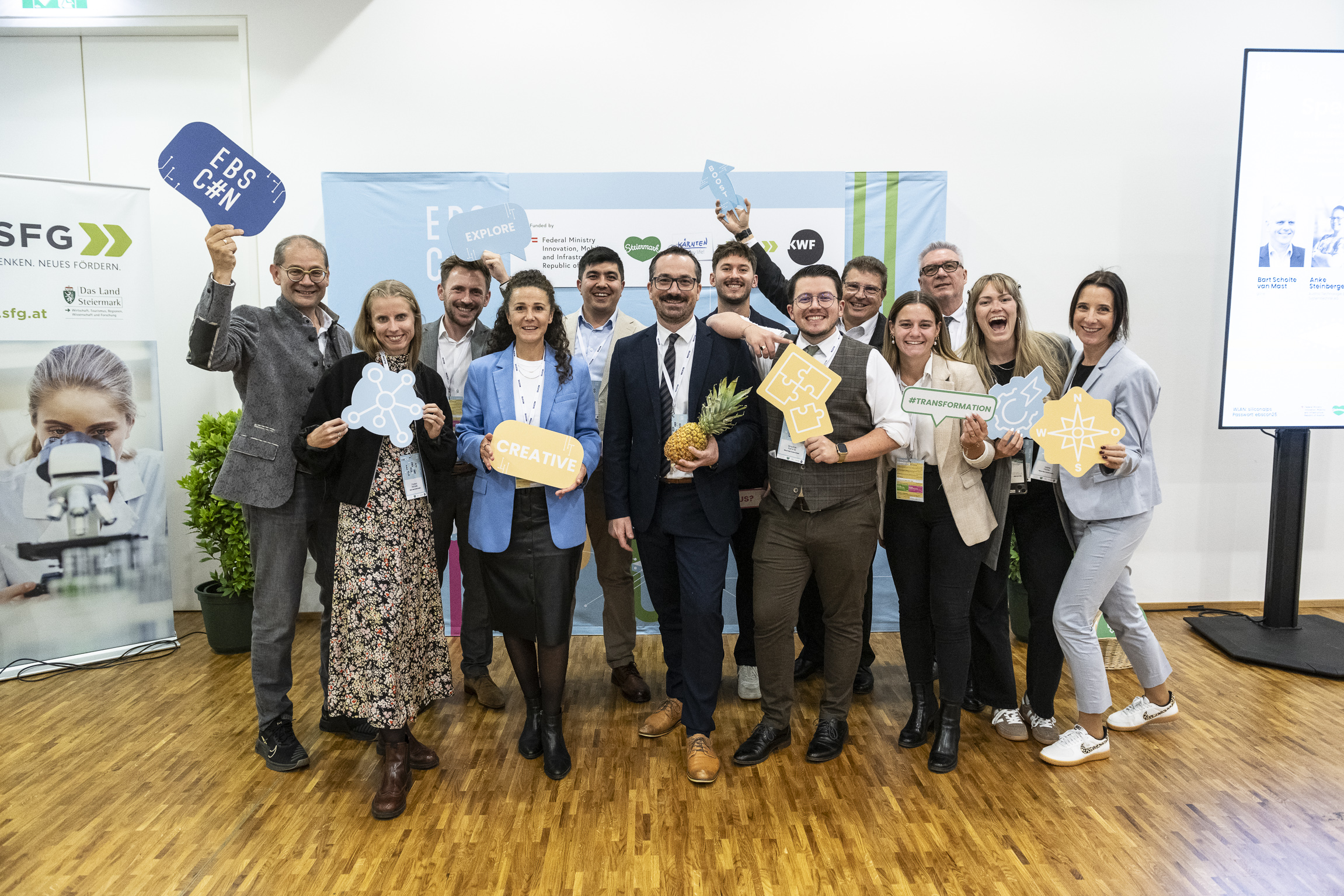 Eine Gruppe beim „Hummelnest Start-up Accelerator Graz“ posiert in einem Innenraum, lächelt und hält motivierende Schilder und Symbole in die Kamera; eine Person hält eine Ananas in der Hand.