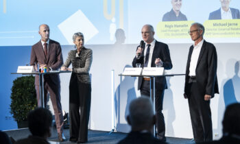 Vier Geschäftsleute sprechen auf der Bühne bei einer Podiumsdiskussion des „Hummelnest Start-up Accelerator Graz“ – mit Mikrofonen und Notizen in der Hand – vor einem blau-weißen Hintergrund.