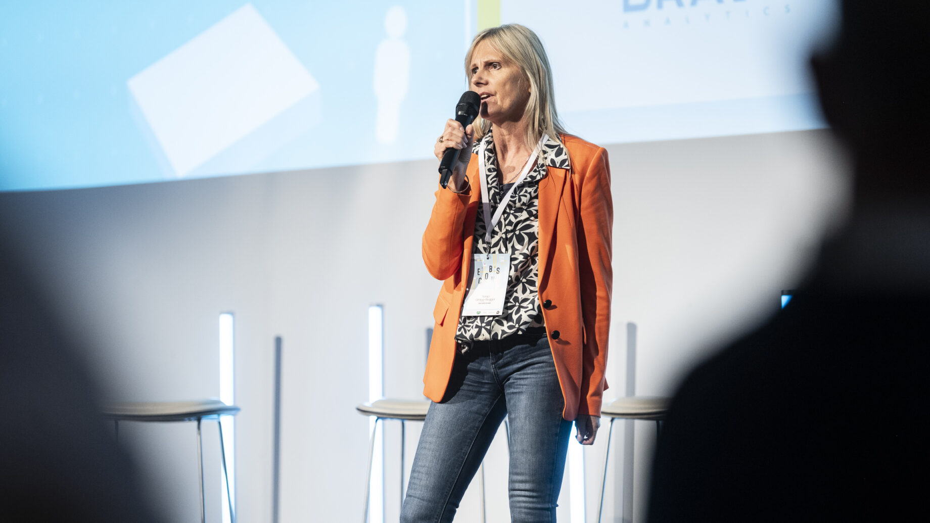 Eine Frau in einem orangefarbenen Blazer hält auf einer Konferenz eine Rede, während im Hintergrund die Logos von „BRAVE“ und dem Start-up-Accelerator Hummelnest Graz zu sehen sind.