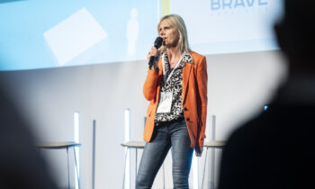 Eine Frau in einem orangefarbenen Blazer hält auf einer Konferenz eine Rede, während im Hintergrund die Logos von „BRAVE“ und dem Start-up-Accelerator Hummelnest Graz zu sehen sind.