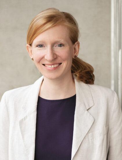 Eine Frau mit roten Haaren, die zu einem Pferdeschwanz gebunden sind, trägt einen hellen Blazer und eine dunkle Bluse und steht mit den Händen in den Taschen vor einem neutralen Hintergrund und lächelt.