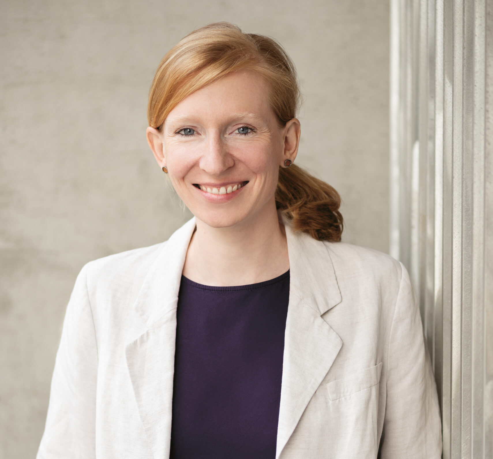 Eine Frau mit roten Haaren, die zu einem Pferdeschwanz gebunden sind, trägt einen hellen Blazer und eine dunkle Bluse und steht mit den Händen in den Taschen vor einem neutralen Hintergrund und lächelt.
