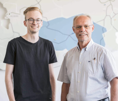 Zwei Männer stehen nebeneinander in einem Raum, lächeln in die Kamera, im Hintergrund ist eine Karte an der Wand zu sehen.