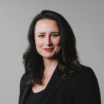 Eine Frau mit langen dunklen Haaren, die einen schwarzen Blazer und ein schwarzes Oberteil trägt und leicht lächelt, posiert vor einem schlichten hellgrauen Hintergrund.