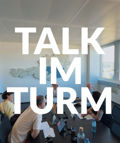 Fünf Personen sitzen um einen modernen Bürotisch herum. Der Text lautet: „TALK IM TURM"