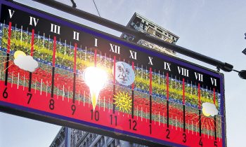 Über einer Straßenecke in der Stadt, an der sich hohe Gebäude befinden und unter der ein Schild mit der Aufschrift „Mercer St.“ zu sehen ist, hängt ein farbenfrohes Sonnenuhrschild mit Zahlen und Wettersymbolen.
