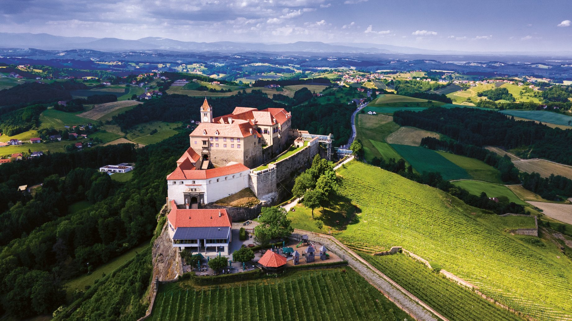 Eine Burg auf einem Hügel mit roten Dächern, umgeben von Feldern, Wäldern und fernen Hügeln unter einem blauen Himmel mit vereinzelten Wolken.