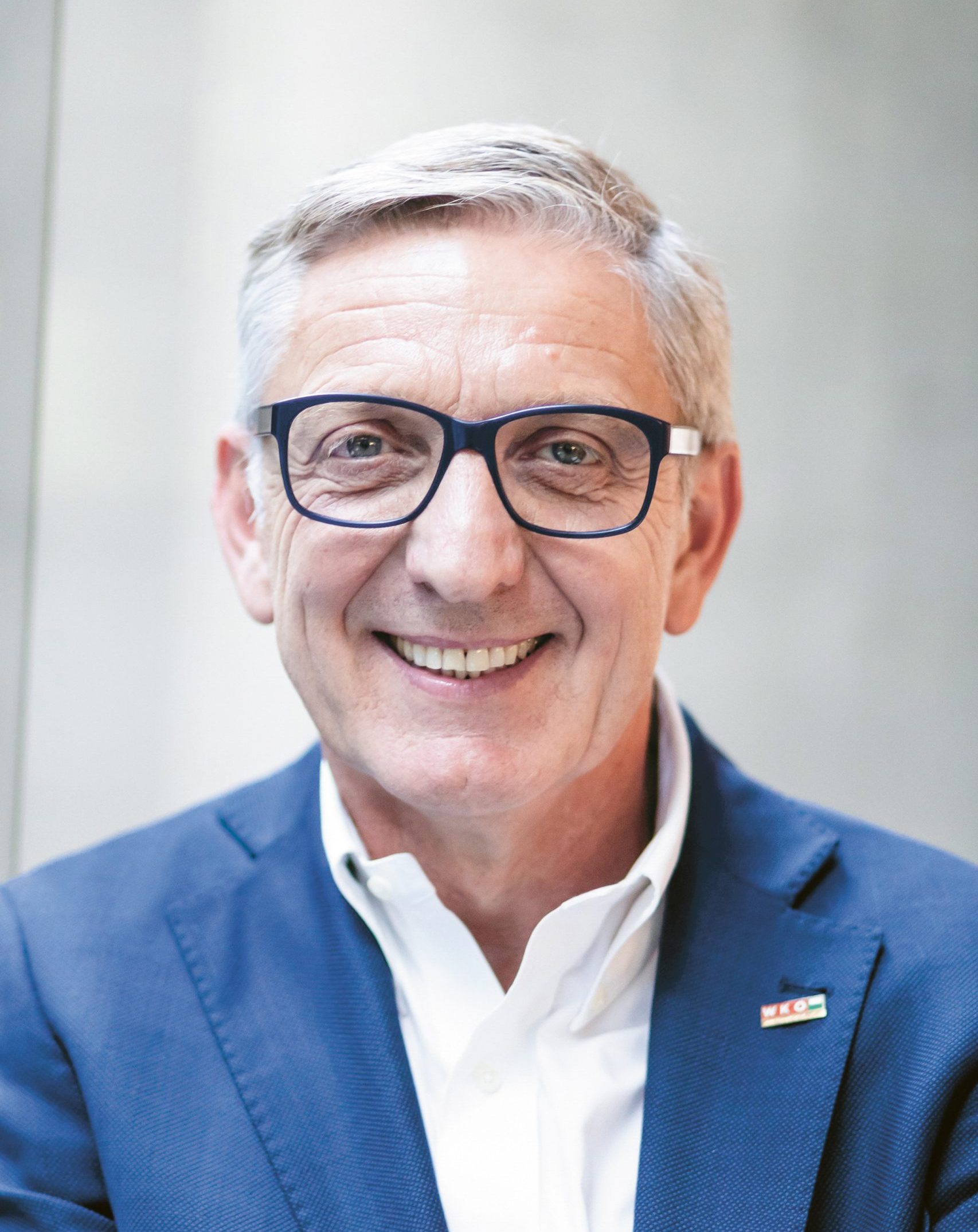 Ein lächelnder älterer Mann mit grauen Haaren und Brille, der einen blauen Anzug und ein weißes Hemd trägt und in einem Raum steht.