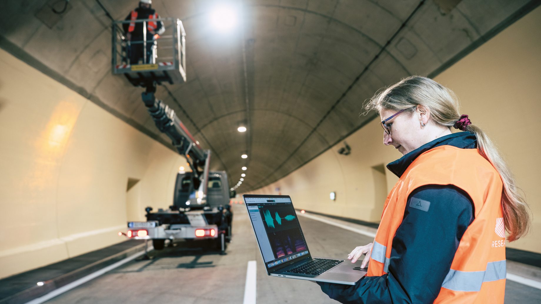 Eine Frau in einer orangefarbenen Warnweste arbeitet an einem Laptop in einem Tunnel, während ein Arbeiter in einem Aufzug die Tunneldecke inspiziert.