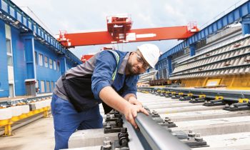 Ein Arbeiter mit Schutzhelm und Schutzkleidung inspiziert Eisenbahnschienen auf einem Industriegelände mit großen Kränen und blauen Gebäuden.
