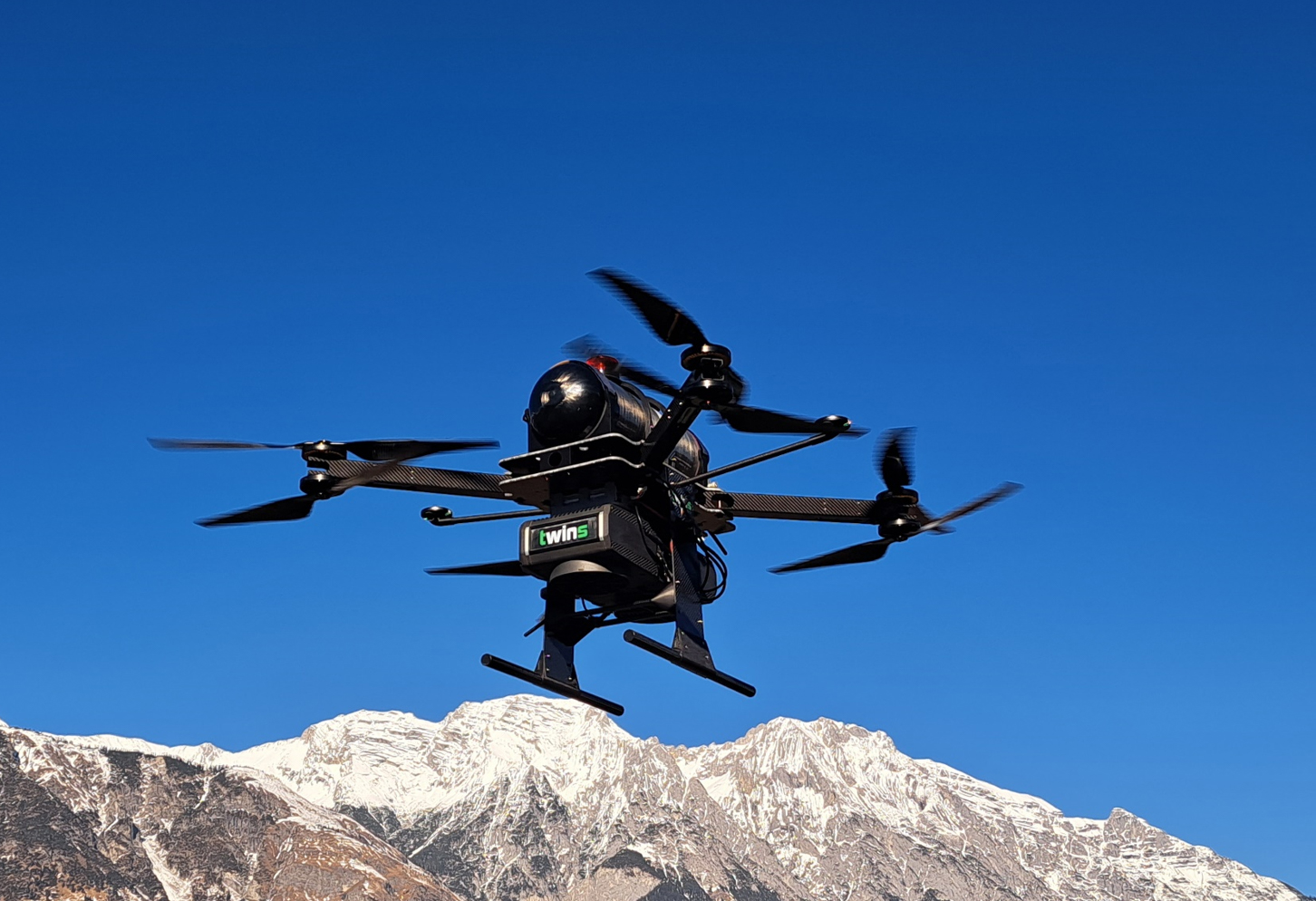 Eine schwarze Drohne mit vier Rotoren fliegt vor schneebedeckten Bergen unter einem strahlend blauen Himmel.