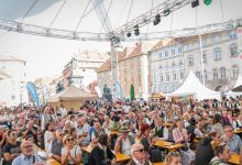 Bei einer Freiluftveranstaltung auf einem europäischen Stadtplatz, umgeben von historischen Gebäuden, sitzt eine große Menschenmenge an langen Tischen unter einem Zelt.
