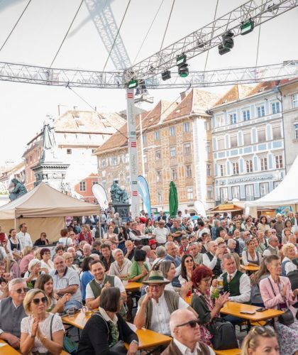 Bei einer Freiluftveranstaltung auf einem europäischen Stadtplatz, umgeben von historischen Gebäuden, sitzt eine große Menschenmenge an langen Tischen unter einem Zelt.