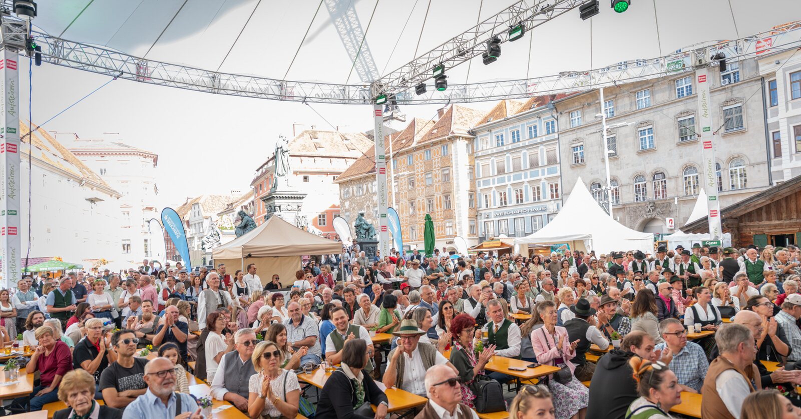 Bei einer Freiluftveranstaltung auf einem europäischen Stadtplatz, umgeben von historischen Gebäuden, sitzt eine große Menschenmenge an langen Tischen unter einem Zelt.