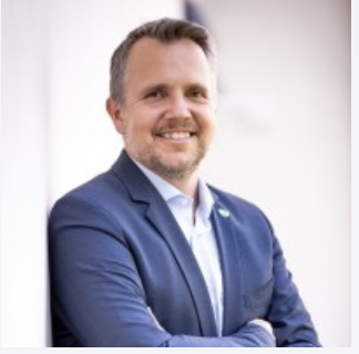 Ein Mann in einem blauen Sakko steht mit verschränkten Armen lächelnd vor einer weißen Wand in einer hell beleuchteten Umgebung und ist bereit für „Aufsteirern und Maxlaunmarkt“.