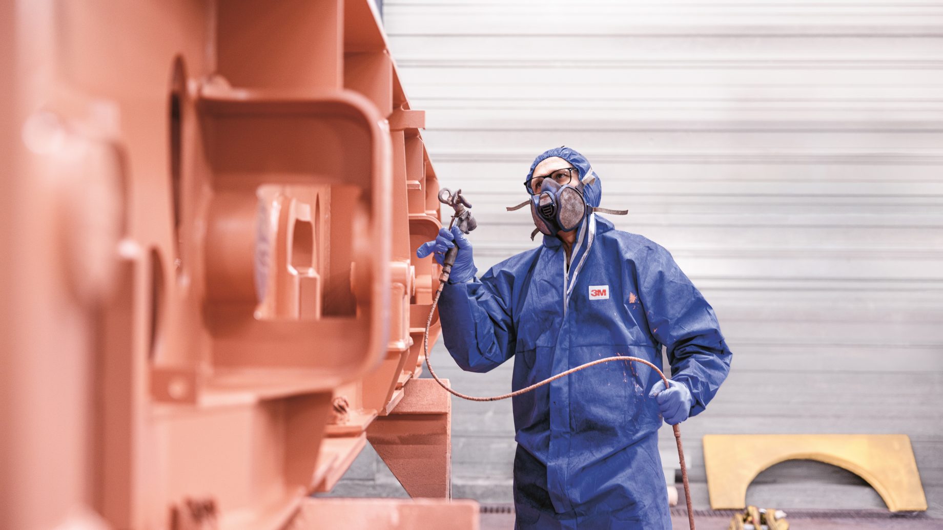 Eine Person in Schutzkleidung und mit Maske besprüht in einem Innenraum eine große Metallkonstruktion mit Sprühfarbe.