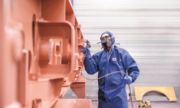 Eine Person in Schutzkleidung und mit Maske besprüht in einem Innenraum eine große Metallkonstruktion mit Sprühfarbe.