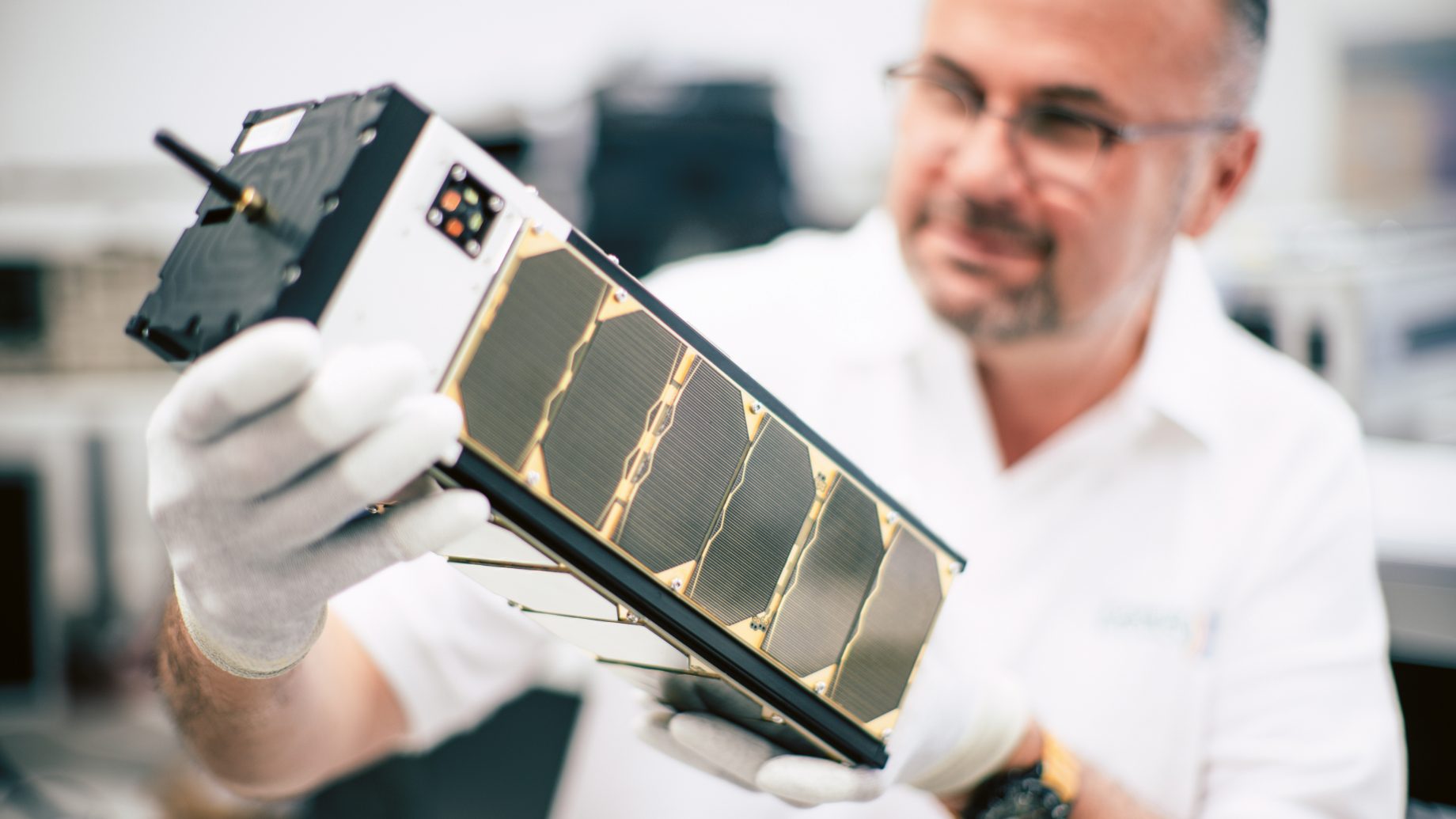Eine Person mit weißen Handschuhen hält einen kleinen Satelliten in der Hand und untersucht ihn in einem Laborraum genau.