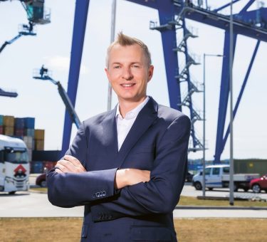 Ein lächelnder Mann im Anzug steht mit verschränkten Armen an einem geschäftigen Hafen; im Hintergrund sind große Kräne, Lastwagen und Schiffscontainer zu sehen.