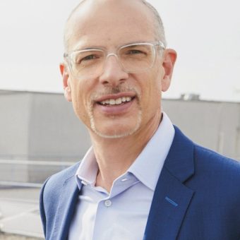 Ein kahlköpfiger Mann mit Brille, einem leichten Schnurrbart und einem Ziegenbart, der ein blaues Sakko und ein helles Hemd trägt und im Freien lächelt.