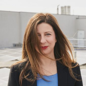Eine Frau mit langen braunen Haaren steht im Freien, trägt einen schwarzen Blazer und ein blaues Oberteil und lächelt, wobei ihr Haar ihr Gesicht teilweise verdeckt.
