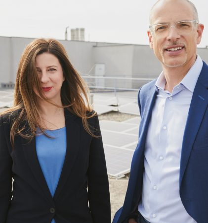 Zwei Personen in Geschäftskleidung stehen auf einem Dach mit Solarmodulen und lächeln in die Kamera. Der Himmel ist klar und das Gebäude ist modern.