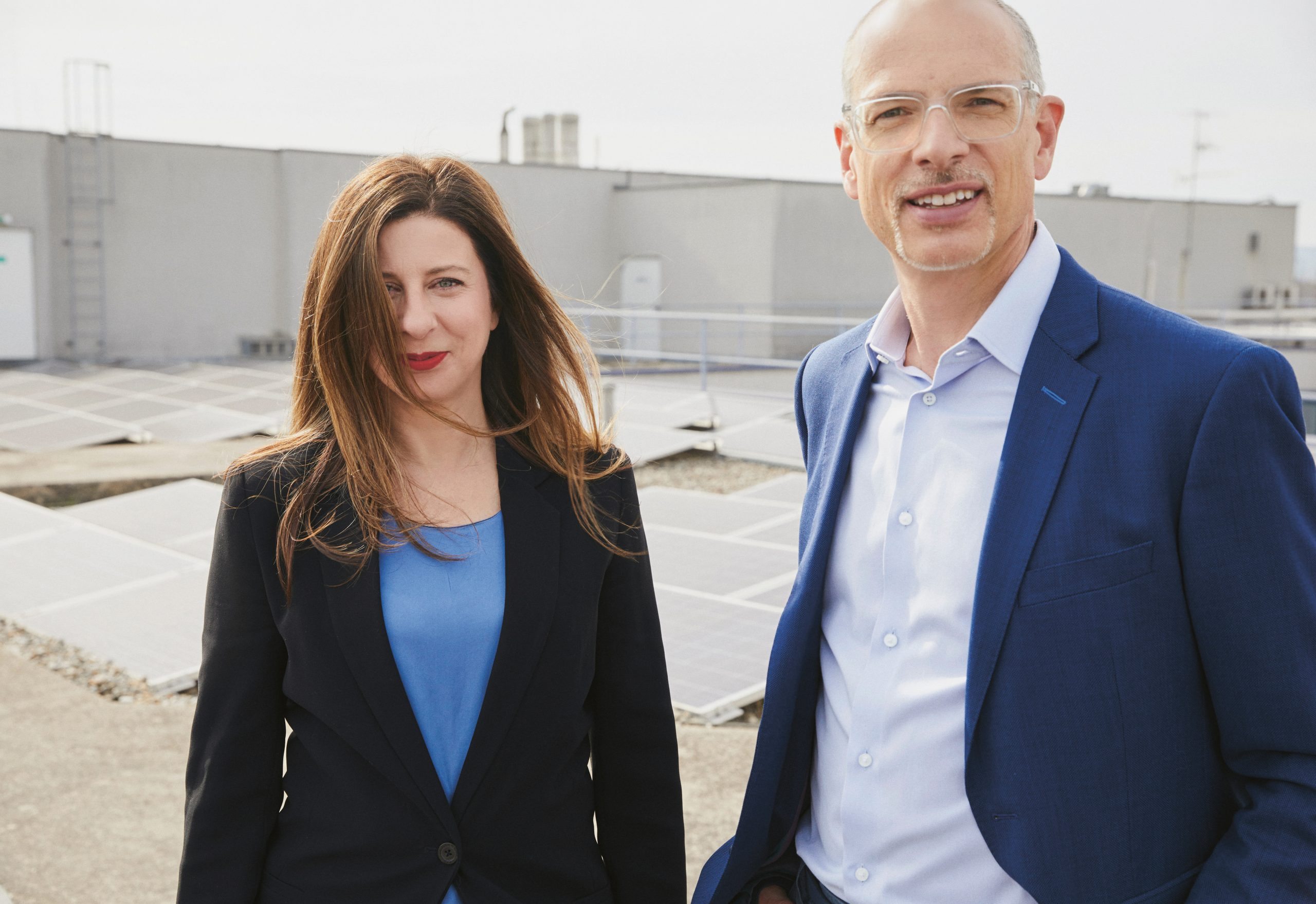 Zwei Personen in Geschäftskleidung stehen auf einem Dach mit Solarmodulen und lächeln in die Kamera. Der Himmel ist klar und das Gebäude ist modern.