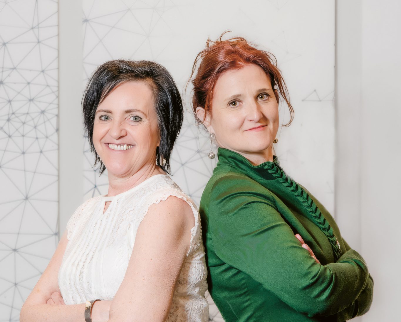 Zwei Frauen stehen Rücken an Rücken, die Arme verschränkt, und lächeln; eine trägt ein weißes Oberteil, die andere eine grüne Jacke, vor einem hellen geometrischen Hintergrund.