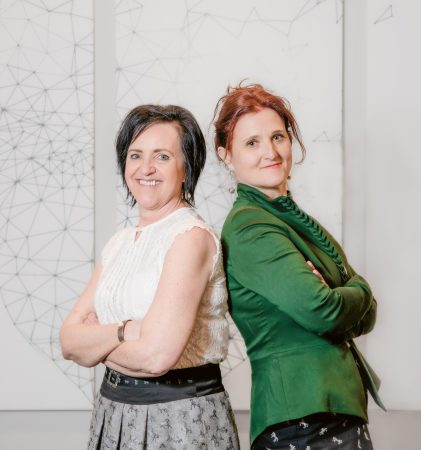 Zwei Frauen stehen Rücken an Rücken und lächeln; die eine trägt eine weiße Bluse und einen Rock, die andere einen grünen Blazer, hinter ihnen ist eine Wandgestaltung mit geometrischen Motiven zu sehen.