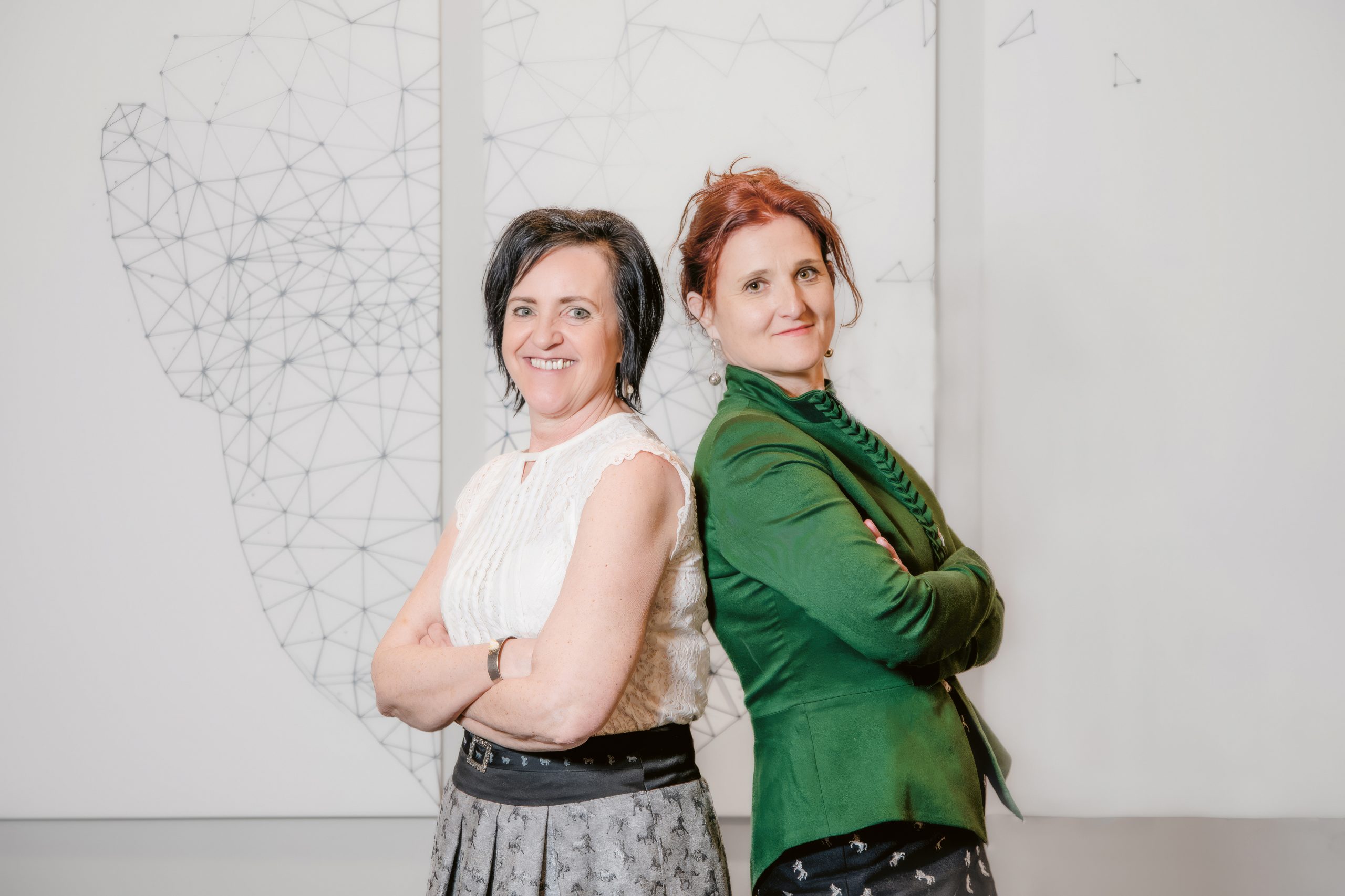 Zwei Frauen stehen Rücken an Rücken und lächeln; die eine trägt eine weiße Bluse und einen Rock, die andere einen grünen Blazer, hinter ihnen ist eine Wandgestaltung mit geometrischen Motiven zu sehen.