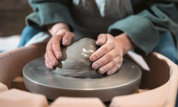 Nahaufnahme einer Person, die nassen Ton auf einer Töpferscheibe formt und dabei mit beiden Händen den sich drehenden Ton führt.