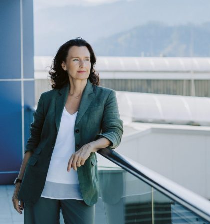 Eine Frau im Anzug steht auf einem Balkon und blickt auf Industriegebäude und einen in der Ferne stehenden Kran, im Hintergrund sind Berge zu sehen.