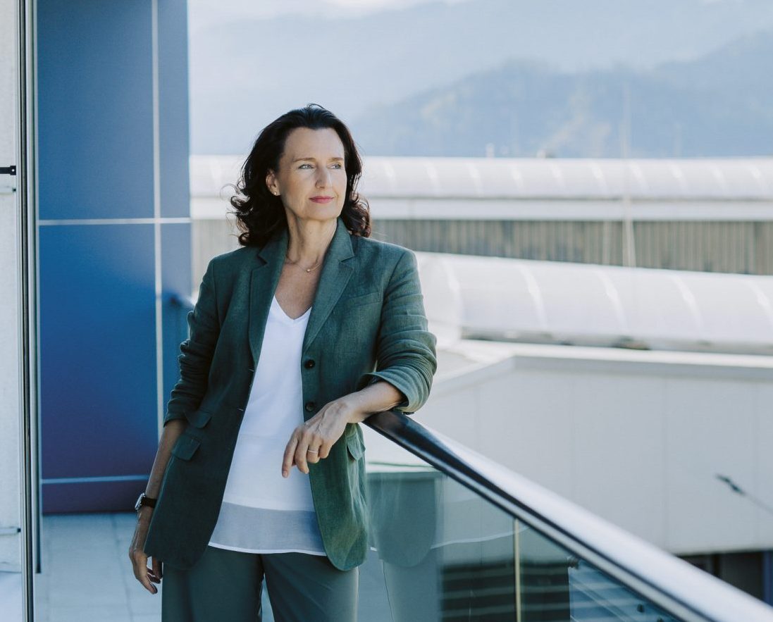 Eine Frau im Anzug steht auf einem Balkon und blickt auf Industriegebäude und einen in der Ferne stehenden Kran, im Hintergrund sind Berge zu sehen.