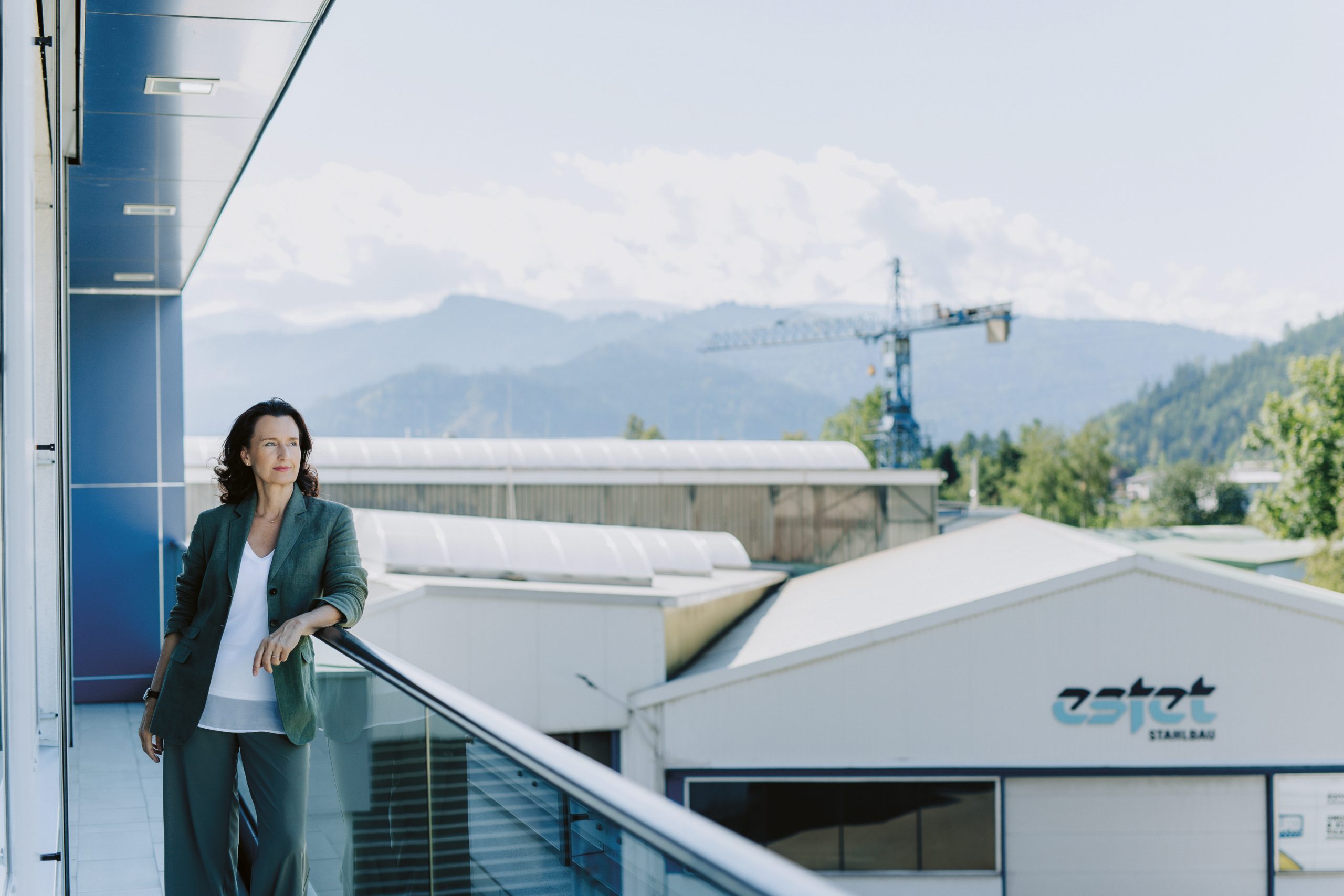 Eine Frau in Business-Kleidung steht auf einem Balkon mit Blick auf Industriegebäude und einen Kran, im Hintergrund sind Berge zu sehen.
