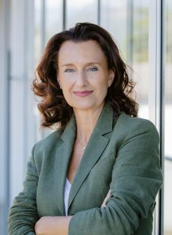 Eine Frau mit welligem braunem Haar und einem grünen Blazer steht drinnen, die Arme verschränkt, und lächelt sanft vor großen Fenstern.