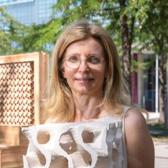 Eine Frau mit blonden Haaren und Brille steht im Freien und hält eine abstrakte weiße Skulptur in der Hand; im Hintergrund sind Bäume und moderne Gebäude zu sehen.