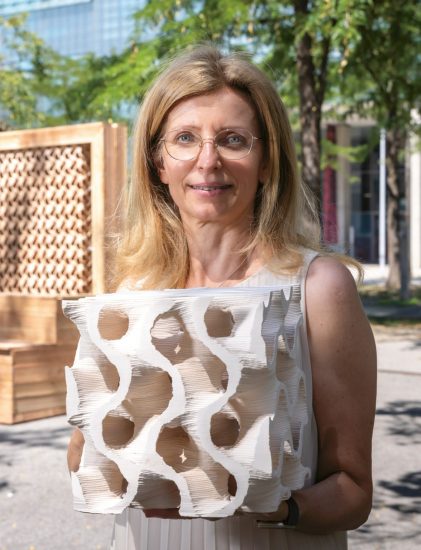 Eine Frau mit Brille hält ein weißes, skulpturales Objekt mit geometrischen Löchern in der Hand und steht im Freien in der Nähe von Bäumen und modernen Holzkonstruktionen.